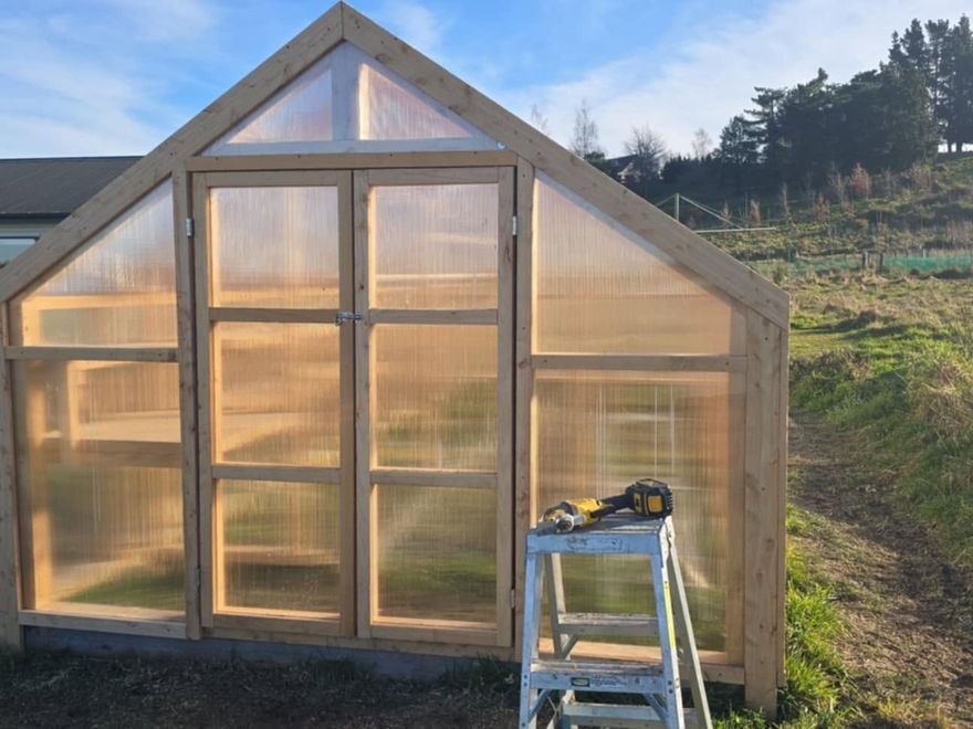 Front of a finished timber and polycarbonate greenhouse with a hinged door, in a paddock beside a house